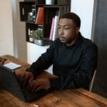 Emprego Remoto com salário a partir de R$ 16.000 (CLT) African American man in black shirt typing on laptop at office desk