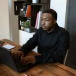 Emprego Remoto com salário a partir de R$ 16.000 (CLT) African American man in black shirt typing on laptop at office desk