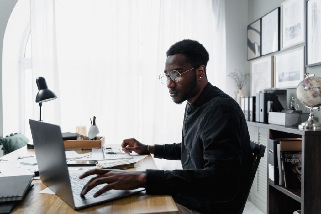 Analista de Marketing Júnior e mais vagas Home Office Black man in glasses working on laptop at office desk, surrounded by documents and modern decor.
