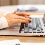 Remotas no Efí Bank, semana de 4 dias e R$ 1.760 de VA/VR A close-up view of hands typing on a laptop keyboard with a focus on manicured nails and technology.