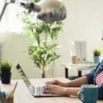 Vagas Remotas no Sicoob, com VR/VA de até R$ 2.300 Woman smiling while working on a laptop in a modern office with plants.