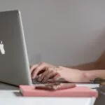 Vagas 100% Remotas na YDUQS Close-up of a woman typing on a laptop at a modern, minimalist desk with a phone nearby.