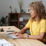 Remotas com salário até R$ 24.000, CLT e PJ A woman working remotely from home, typing on a keyboard at her work desk.