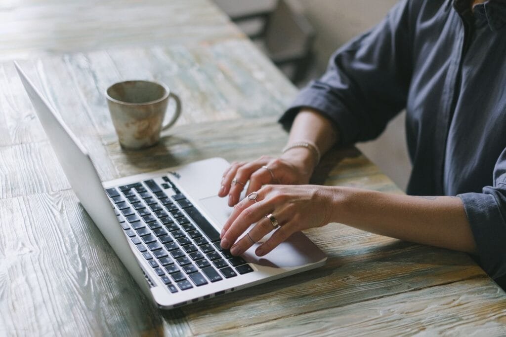 Analista de Relacionamento Júnior e +vagas 100% remotas! Crop faceless female freelancer in casual outfit sitting at table with cup of drink and surfing on laptop in light workspace