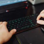 Remotas na Cadastra, com VR de R$ 700,00 Closeup of hands typing on an RGB keyboard in a modern office setting.