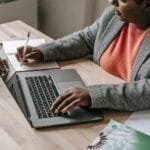 Customer Success Remoto e + vagas com VA/VR de R$ 950 From above of crop anonymous African American female in formal clothes sitting at table with laptop and writing in notebook