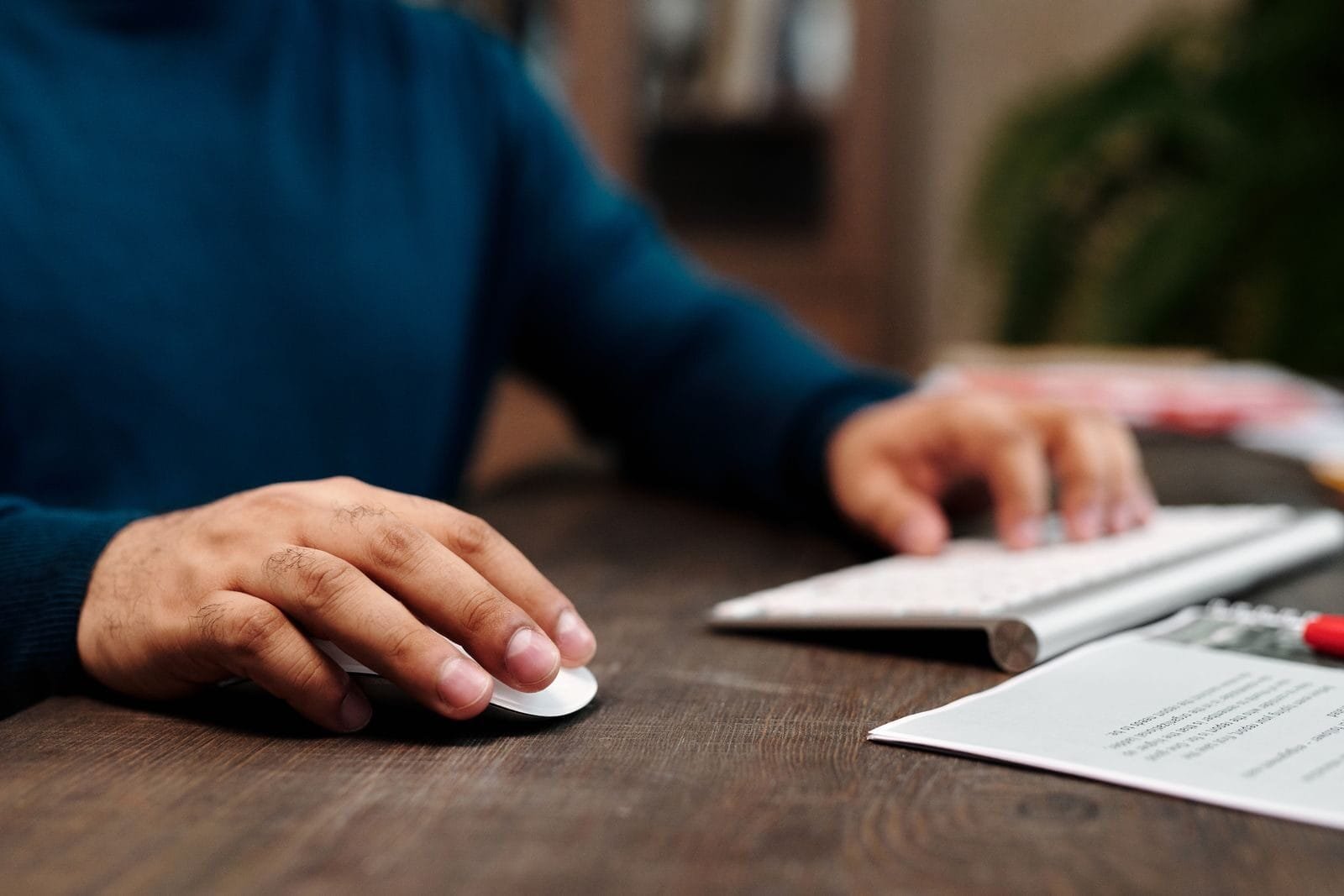 Vagas Remotas com R$ 1.700 em Benefícios Close-up of a man using a wireless mouse and keyboard on a wooden desk.