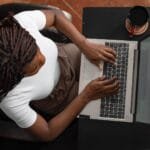 Remotas para Analistas, com VA/VR de R$ 700,00 Overhead shot of a woman typing on a laptop at home, representing remote work.