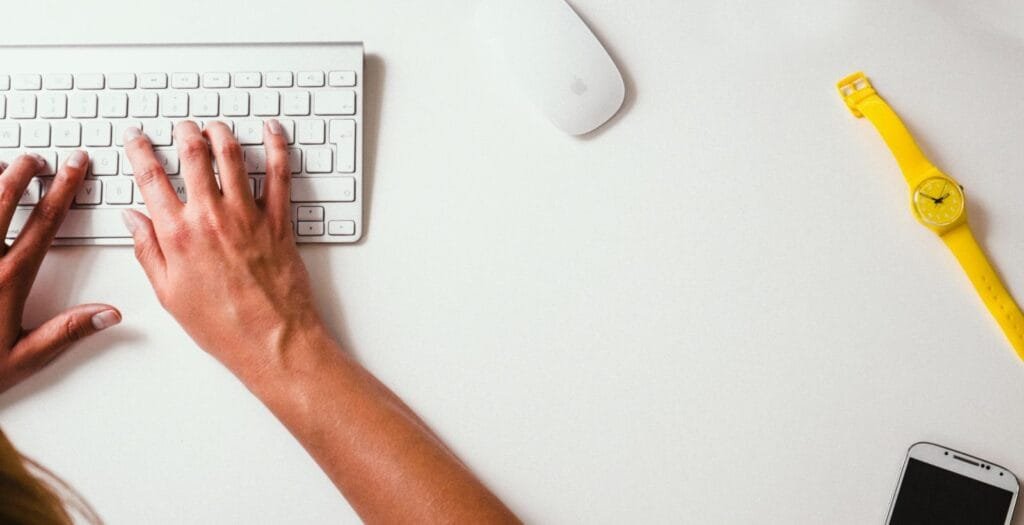 Estágio Financeiro e mais vagas 100% Home Office Close-up of hands typing on a keyboard with a yellow watch on the desk