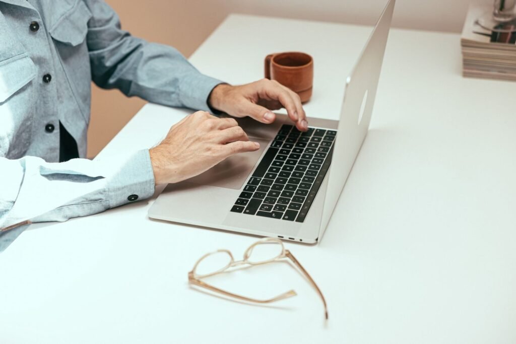 Agente de Atendimento e mais vagas 100% remotas Adult working remotely on laptop with eyeglasses and cup on a white table.
