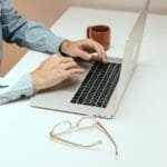 Agente de Atendimento e mais vagas 100% remotas Adult working remotely on laptop with eyeglasses and cup on a white table.