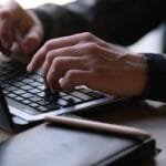 Remotas na Ícaro Tech com R$ 1.000 de VA/VR Close-up photo of hands typing on a laptop, perfect for remote work or digital nomad themes.