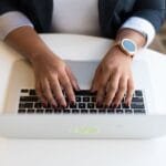 Vagas Remotas com R$ 2.400,00 para posto de trabalho e + benefícios! Woman's hands typing on a wireless laptop at a modern office desk, highlighting technology in business.