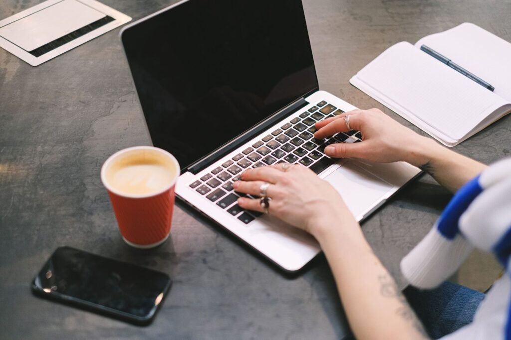Remotas com VR/VA de R$ 837,00 A freelancer typing on a laptop with a cup of coffee, smartphone, and notepad on the table.