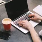 Remotas com VR/VA de R$ 837,00 A freelancer typing on a laptop with a cup of coffee, smartphone, and notepad on the table.