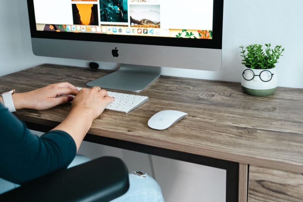 Analista de Recursos Humanos e + vagas 100% remotas! A woman's hands typing on a keyboard at a wooden desk with a computer setup.