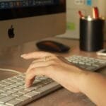 Analista e +vagas remotas com R$ 1.012,50 de VA/VR A close-up of a woman's hand typing on an Apple keyboard in an office setting.