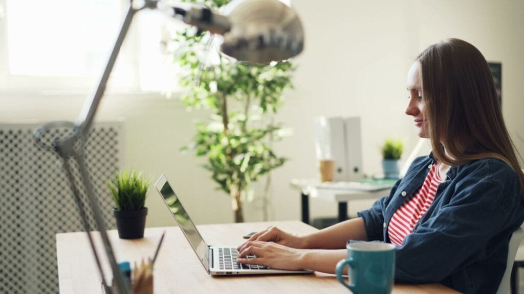Vagas Remotas com R$ 1.200 de VA/VR e mais Benefícios Woman smiling while working on a laptop in a modern office with plants.