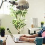 Vagas Remotas com R$ 1.200 de VA/VR e mais Benefícios Woman smiling while working on a laptop in a modern office with plants.