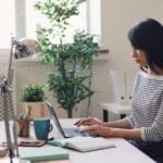 Atendimento Júnior e mais vagas 100% remotas! Focused young woman working on a laptop in a bright office setting with plants and desk accessories.