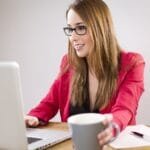 Estágio e Efetiva em vagas 100% Home Office! A smiling young businesswoman in a red jacket works on her laptop while holding a coffee cup.