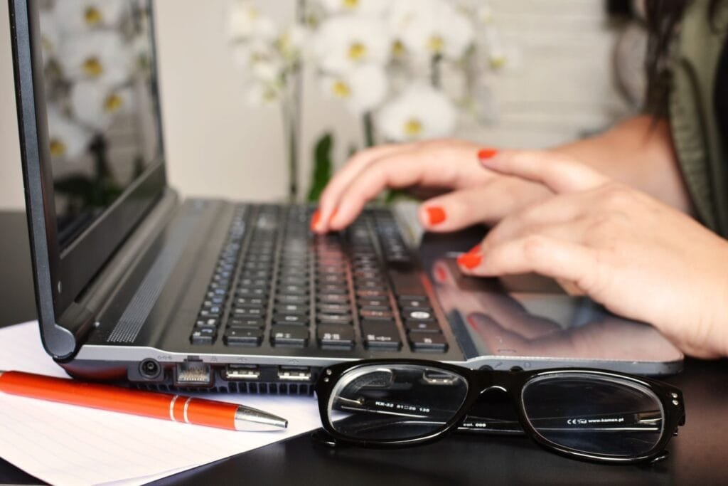 Vagas 100% Remotas na SysMap Close-up of a woman's hands typing on a laptop with glasses and a notepad on the desk.