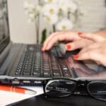 Vagas 100% Remotas na SysMap Close-up of a woman's hands typing on a laptop with glasses and a notepad on the desk.