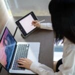 Teletrabalho com VR/VA até R$ 1.500 Asian woman multitasking with laptop and tablet in a modern home office in Bangkok.