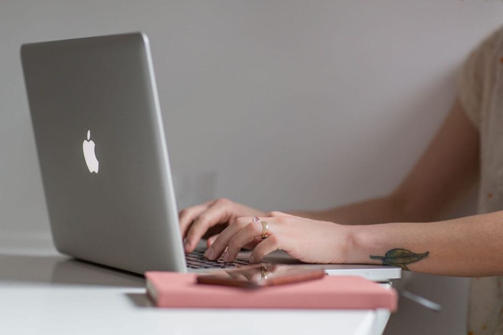 Remotas no QuintoAndar, com VA/VR de R$ 1.000,00 Close-up of a woman typing on a laptop at a modern, minimalist desk with a phone nearby.