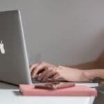 Remotas no QuintoAndar, com VA/VR de R$ 1.000,00 Close-up of a woman typing on a laptop at a modern, minimalist desk with a phone nearby.
