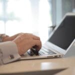 Remotas a Leega com VA de R$ 800,00 Close-up of hands typing on a laptop in a bright, modern office setting.