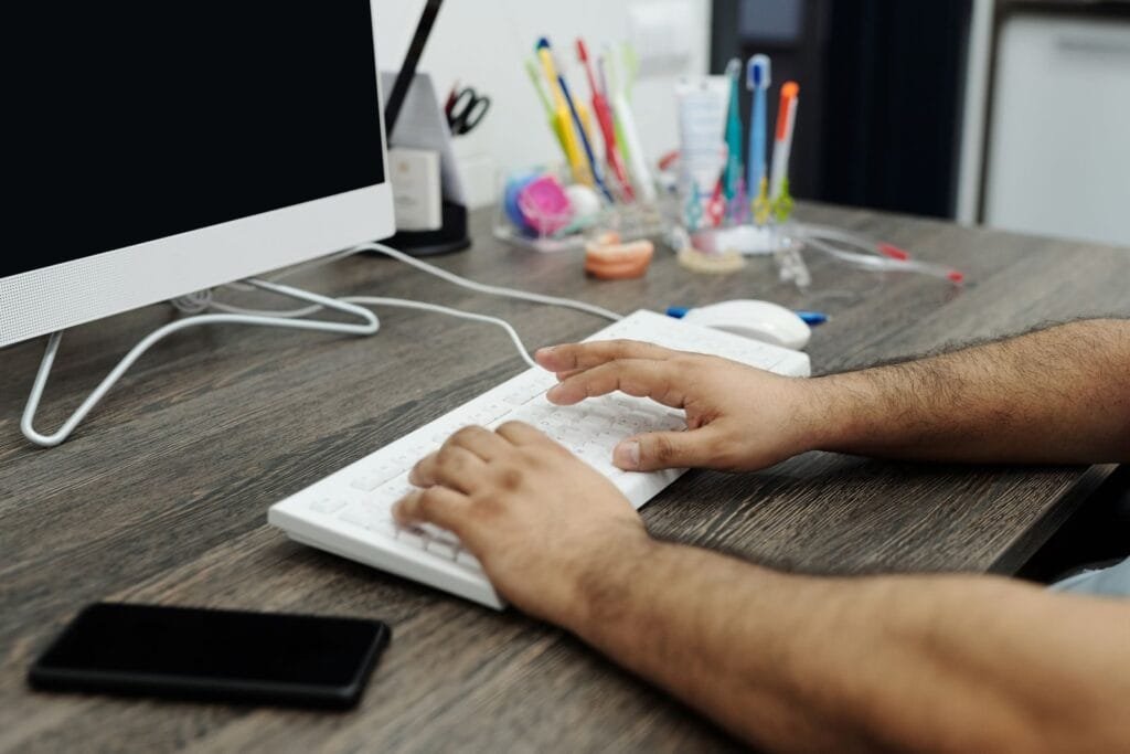 Remotas com salário até R$ 11.000 Close-up of hands typing on a white keyboard at a wooden desk in an office setting with accessories.