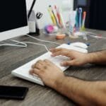 Remotas com salário até R$ 11.000 Close-up of hands typing on a white keyboard at a wooden desk in an office setting with accessories.