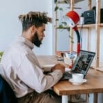 Vagas Remotas com R$ 2.400,00 para posto de trabalho e + benefícios! Side view of African American male freelancer in casual outfit sitting at wooden table and working on laptop near cup of coffee and lamp near shelves in light room