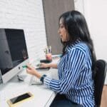 Supervisora de Atendimento, VA/VR médio de R$ 880, e mais Remotas Woman in a striped shirt working at a computer desk in an office setting.