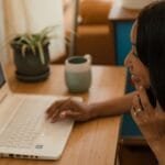 Web Designer e mais vagas 100% remotas! Smiling woman multitasking on phone and laptop at a wooden desk indoors.