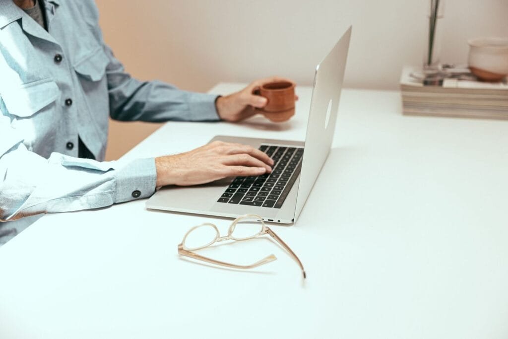 Analista de BI e mais Remotas com VA/VR de R$ 700,00 Casual workspace featuring laptop, coffee cup, and eyeglasses on a white table.