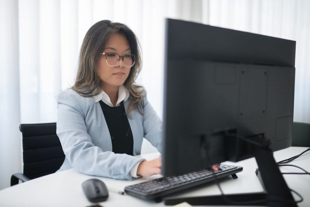 Analista de Produtos e +Vagas Remotas com R$ 1.700 de VR/VA Confident businesswoman focused on work at desk with computer in modern office.