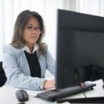 Analista de Produtos e +Vagas Remotas com R$ 1.700 de VR/VA Confident businesswoman focused on work at desk with computer in modern office.