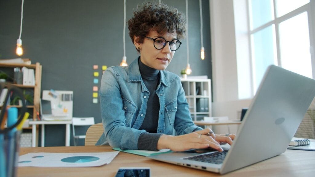 Supervisor de Atendimento e +, com R$ 900,00 de VA/VR A businesswoman using a laptop in a stylish, modern office environment with natural light.