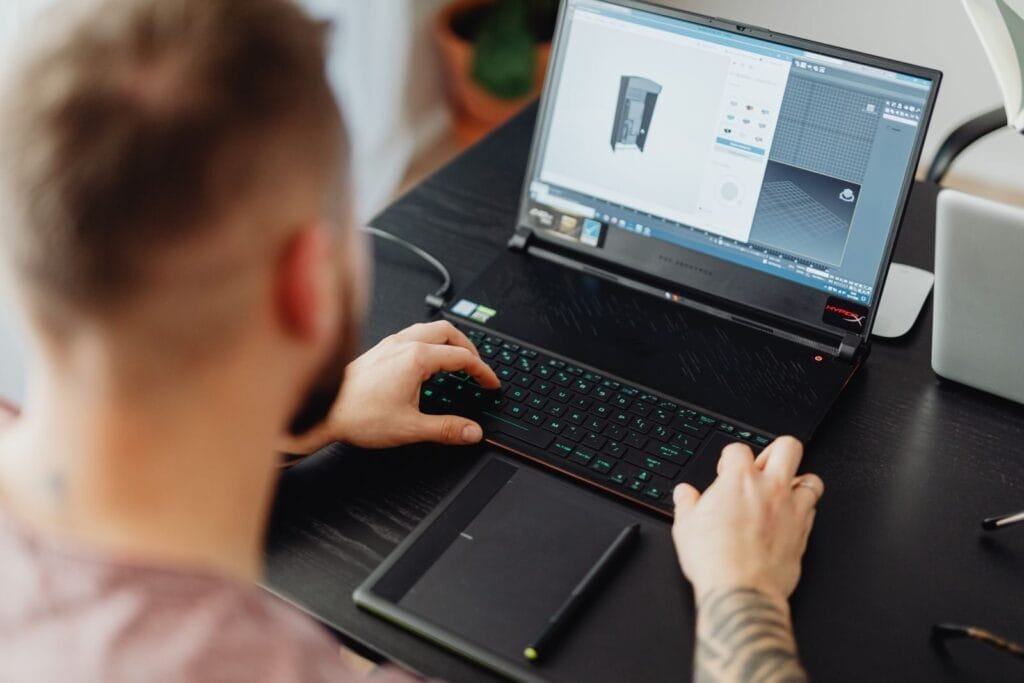 Vagas 100% Remotas na Wicomm Man using laptop and tablet for 3D design, showcasing modern digital work environment.