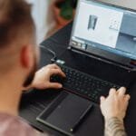Vagas 100% Remotas na Wicomm Man using laptop and tablet for 3D design, showcasing modern digital work environment.