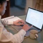 Assistente Comercial e + Remotas com R$ 932 de VR/VA Woman working remotely on laptop with documents and coffee mug on desk.