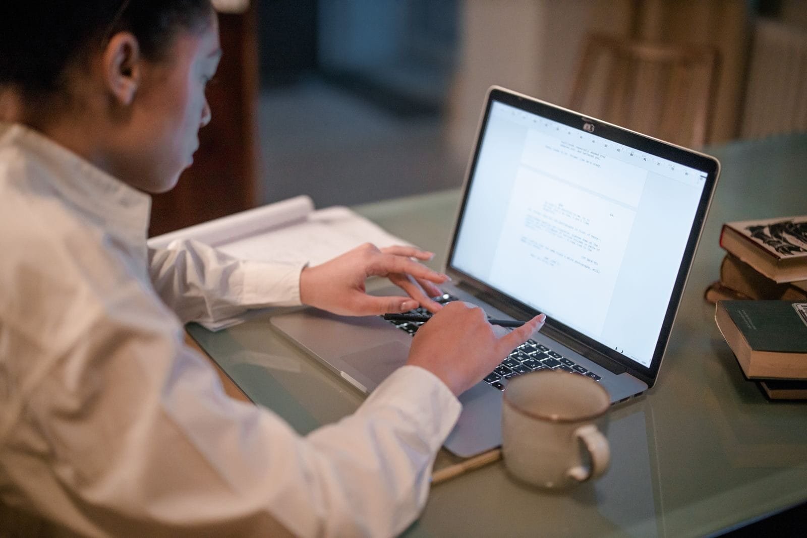 Assistente Comercial e + Remotas com R$ 932 de VR/VA Woman working remotely on laptop with documents and coffee mug on desk.