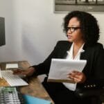 Assistente Fiscal em mais vagas remotas com VR de R$ 922,50 Focused woman working on computer, holding documents, and wearing glasses.