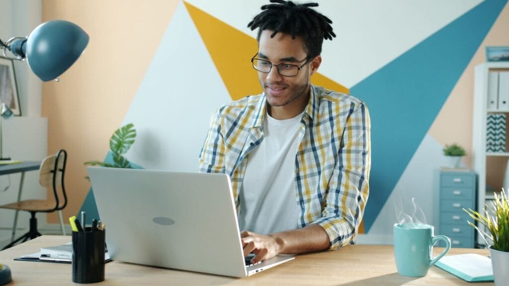 Técnico(a) de Suporte com R$ 950 de VR/VA e +Remotas Young man in a stylish office using a laptop, embodying modern work environment.