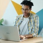 Técnico(a) de Suporte com R$ 950 de VR/VA e +Remotas Young man in a stylish office using a laptop, embodying modern work environment.