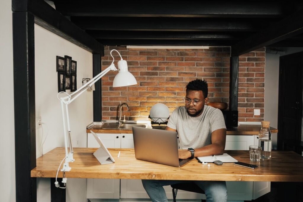 Auxiliar de Vendas e mais vagas 100% remotas! A man working from home at a desk with laptop, lamp, and workspace essentials.