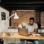 Auxiliar de Vendas e mais vagas 100% remotas! A man working from home at a desk with laptop, lamp, and workspace essentials.