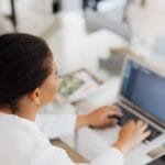 Customer Success com R$ 1.000 de VA/VR e mais Remotas! Black woman working on a laptop in a modern indoor office environment.